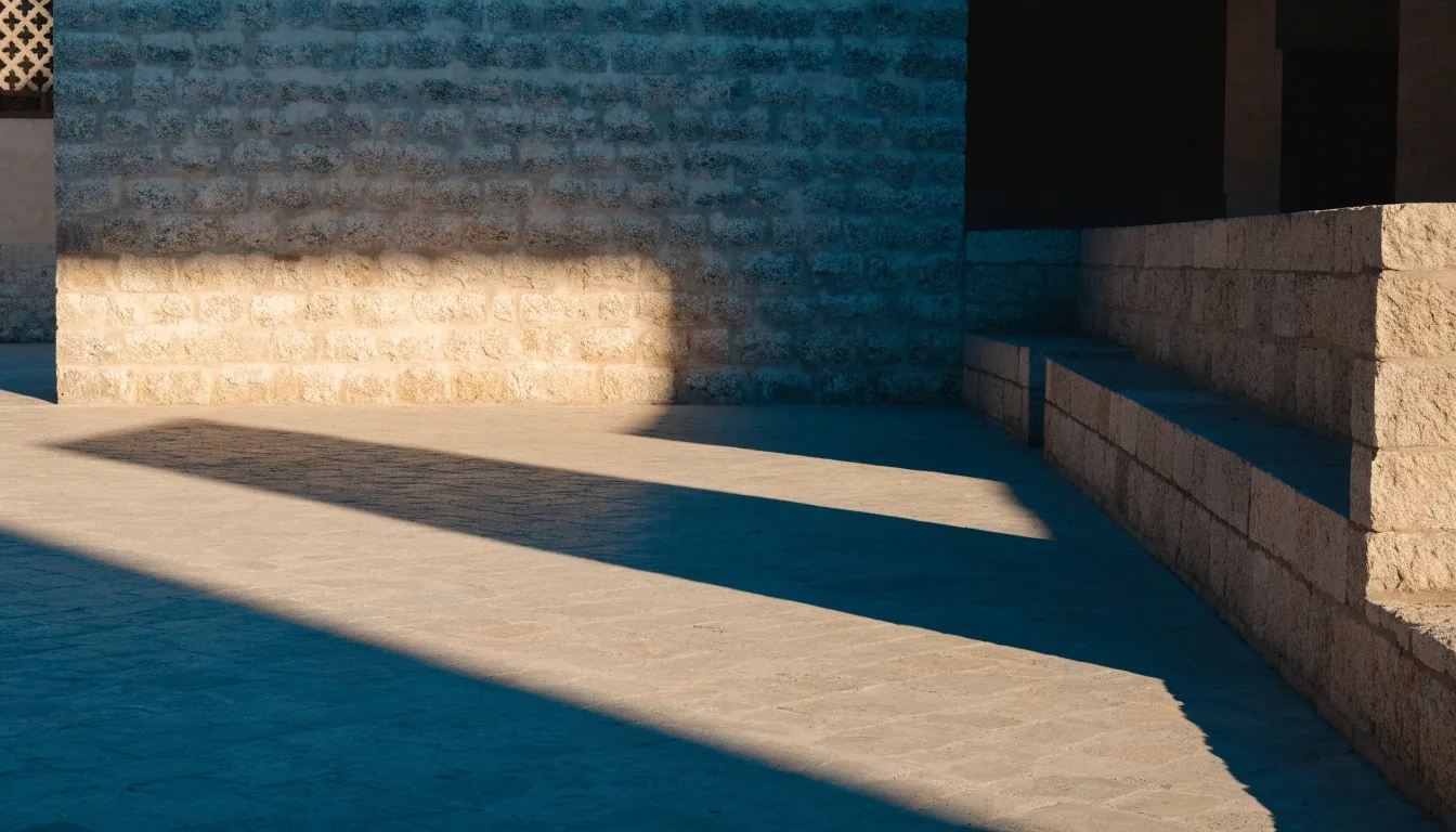 Museum courtyard edge with a cool shadowed stone wall meeting a sunlit paved floor and an empty seating block